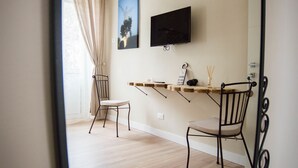 A room with a wooden floor, a TV mounted on the wall, two chairs, a table with a sign, and a window with curtains. - Leucos B&B (Potenza)