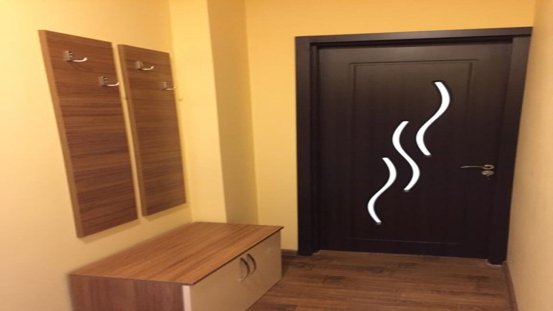 Um corredor com um banco de madeira, uma porta preta com um design abstrato em branco e painéis de madeira na parede.
