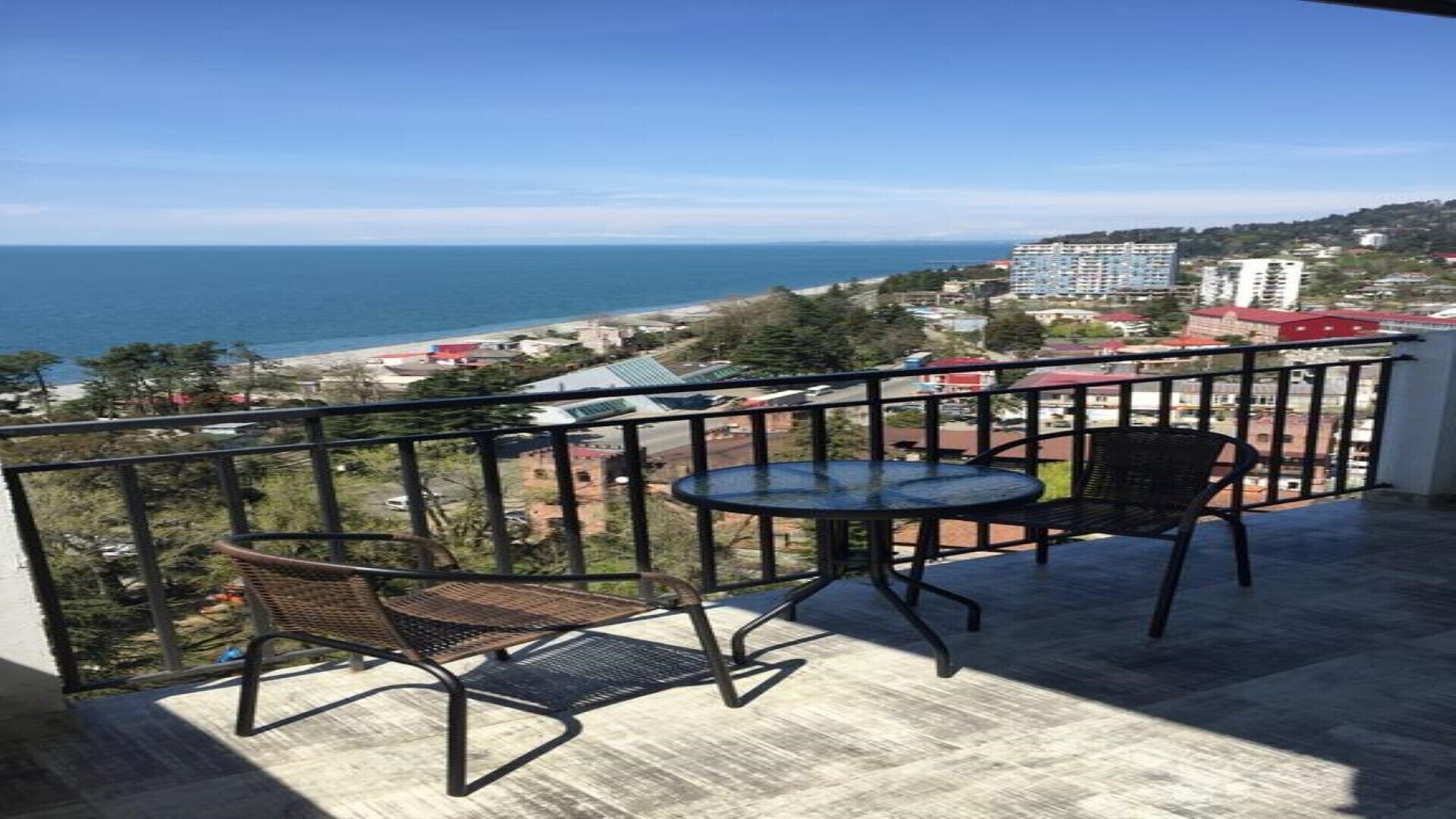 En balkon med to stole og et bord, der giver udsigt over kysten med bygninger og en strand.