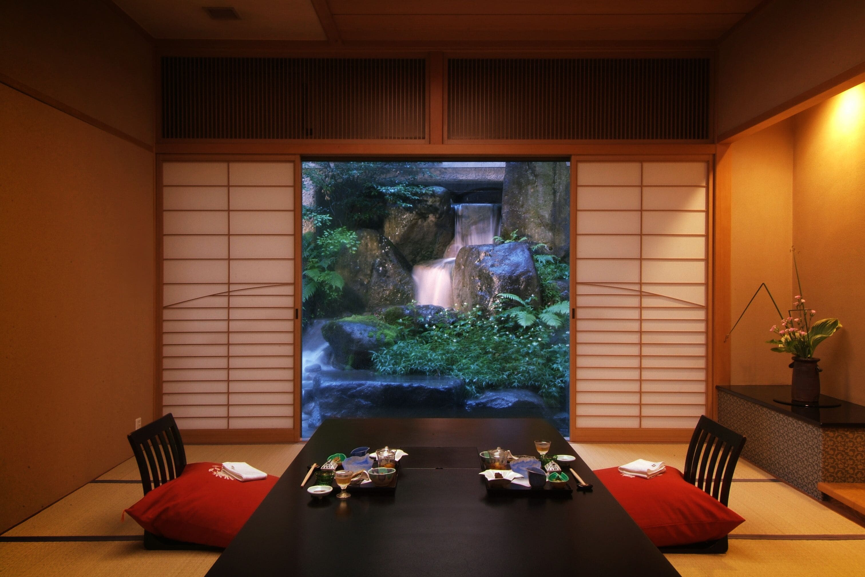 A traditional Japanese room with tatami flooring, a low table set for a meal, and a large sliding door with a waterfall view.