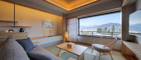 A modern hotel room with a sofa, a wooden table, a chair, a TV, and a large window with a mountain view.