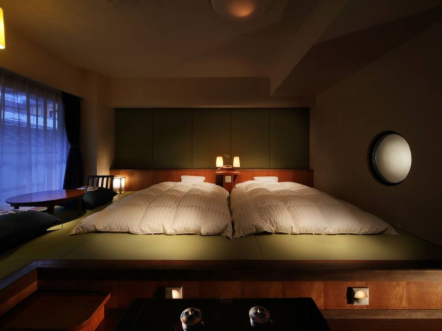 A hotel room with a large bed, two bedside lamps, a round mirror, a wooden headboard, a small table, and a chair.