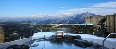 A hot spring with a wooden bench and metal railings, surrounded by snow-covered rocks and a view of mountains.