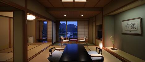Una habitación tradicional japonesa con suelo de tatami, una mesa baja y sillas. Hay un ventanal con vistas a montañas.