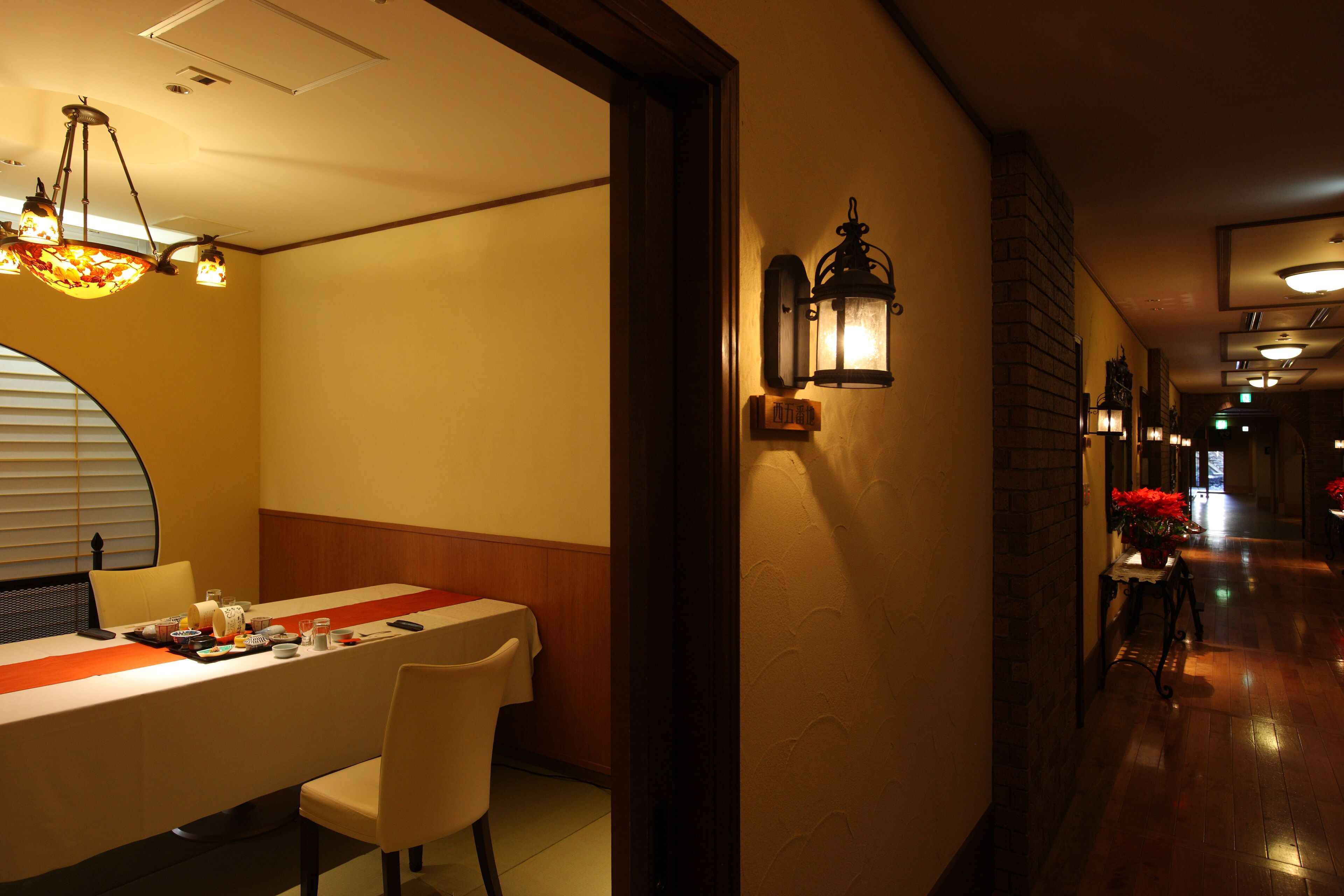 Un comedor de hotel con una mesa larga, sillas y un camino rojo. Se observan faroles en la pared y un candelabro.