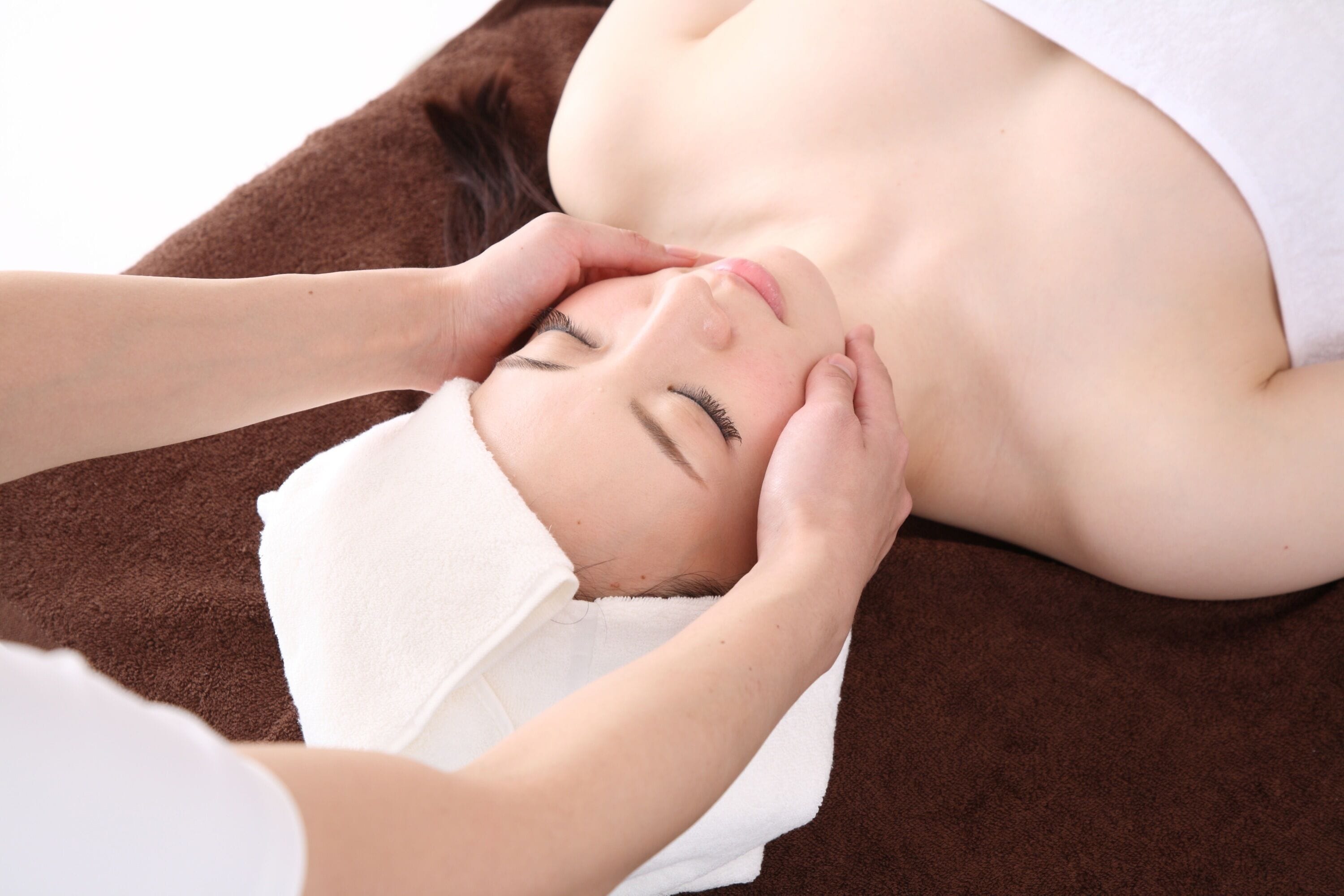A person lying on a brown surface with a white towel, receiving a facial treatment.