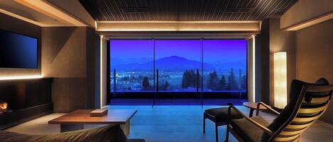 A modern living room with a large window offering a view of a cityscape and mountains at dusk.