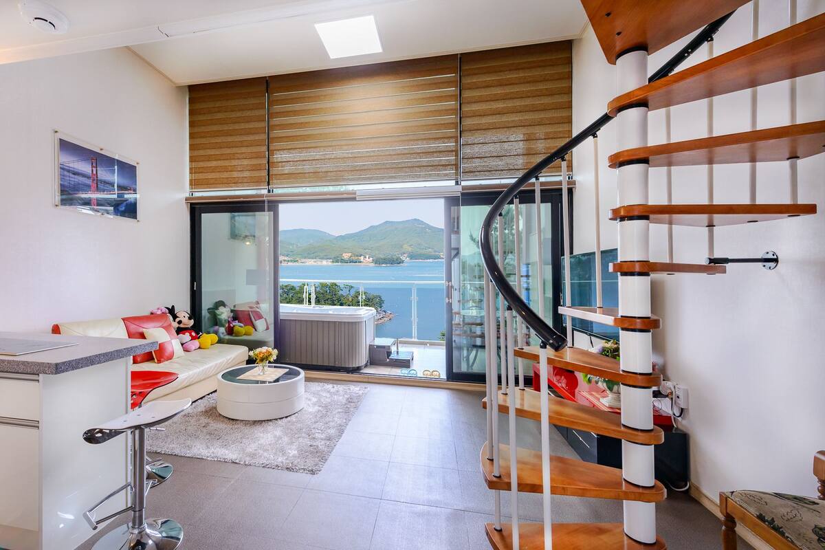 A modern kitchen with a large window, a dining area with a white table and red chairs, and a spiral staircase.