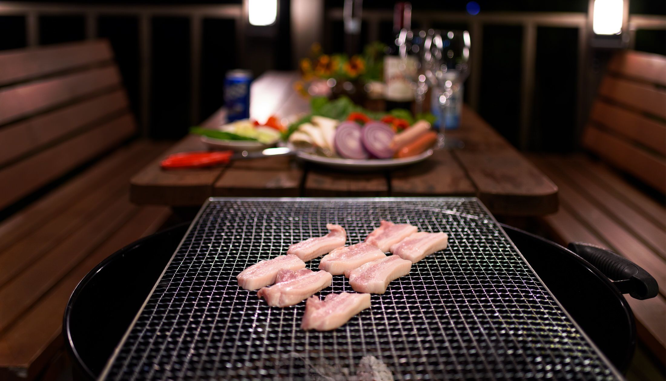 En grill med rått kött i förgrunden och ett bord med grönsaker och tillbehör i bakgrunden.