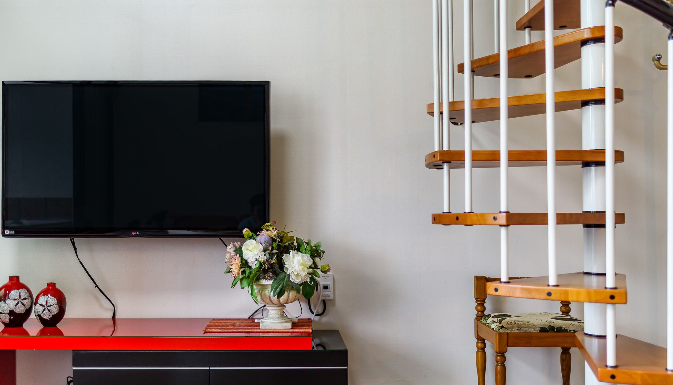 A living room with a flat-screen TV mounted on the wall, a wooden chair, a small table with a vase of flowers, and a red bench.