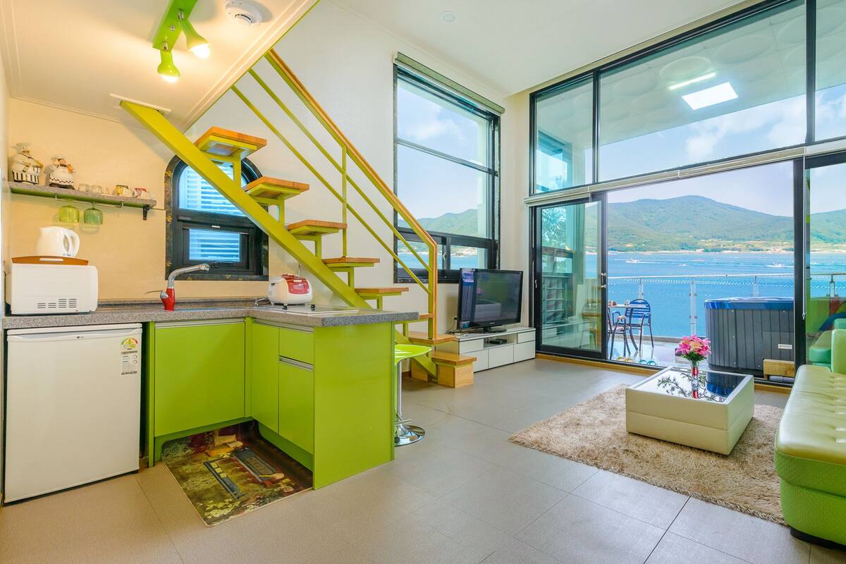 A modern kitchen with a green island, a dining area with a sofa and coffee table, a TV, and a large window with a view of the sea.