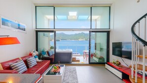 Un salon moderne avec un canapé rouge, une table en verre et un téléviseur. Il est doté d’une grande porte-fenêtre coulissante qui donne sur un balcon offrant une vue sur la mer et les montagnes.