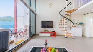 Un salon moderne avec une porte vitrée donnant sur un balcon, un téléviseur fixé au mur, un escalier en colimaçon en bois et une petite table sur laquelle se trouvent une bouteille et des fruits.