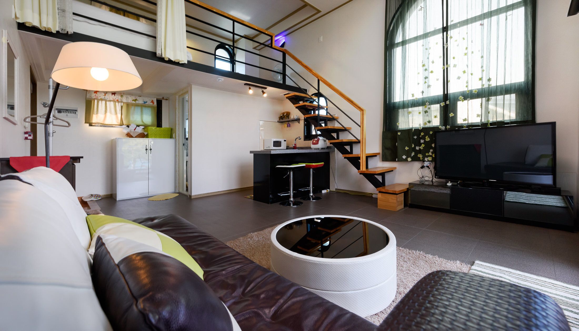 Ein modernes Wohnzimmer mit Sofa, rundem weißen Hocker, Fernseher, Küchenbereich und Treppe.