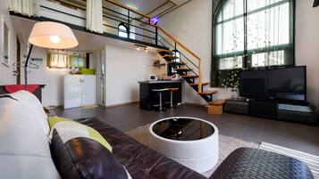 Ein modernes Wohnzimmer mit Sofa, rundem weißen Hocker, Fernseher, Küchenbereich und Treppe.