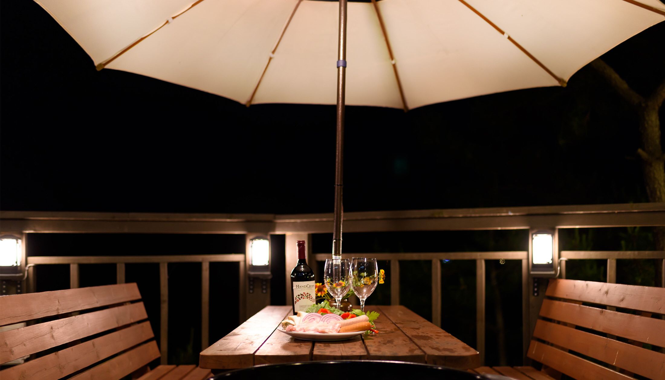 An outdoor dining area with a wooden table, a bottle of wine, and glasses under a large umbrella.