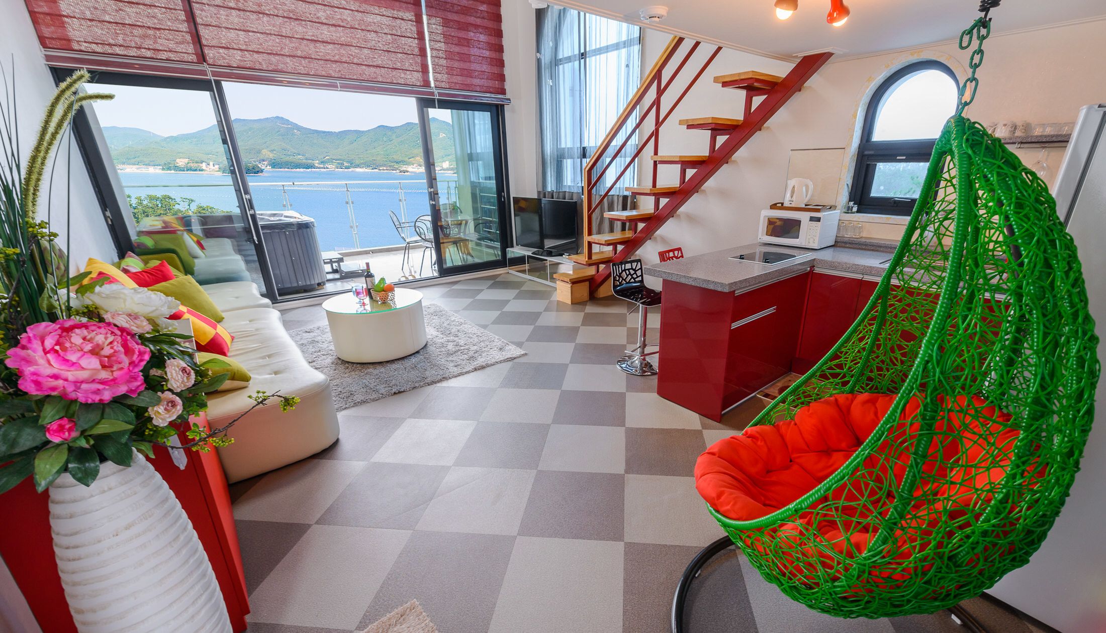 A modern living space with a sofa, a hanging chair, a kitchen area, and a staircase leading to an outdoor balcony with a view of the sea.