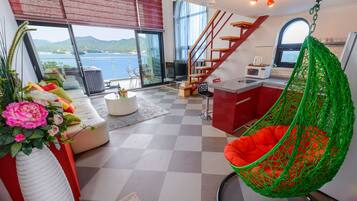 A modern living space with a sofa, a hanging chair, a kitchen area, and a staircase leading to an outdoor balcony with a view of the sea.