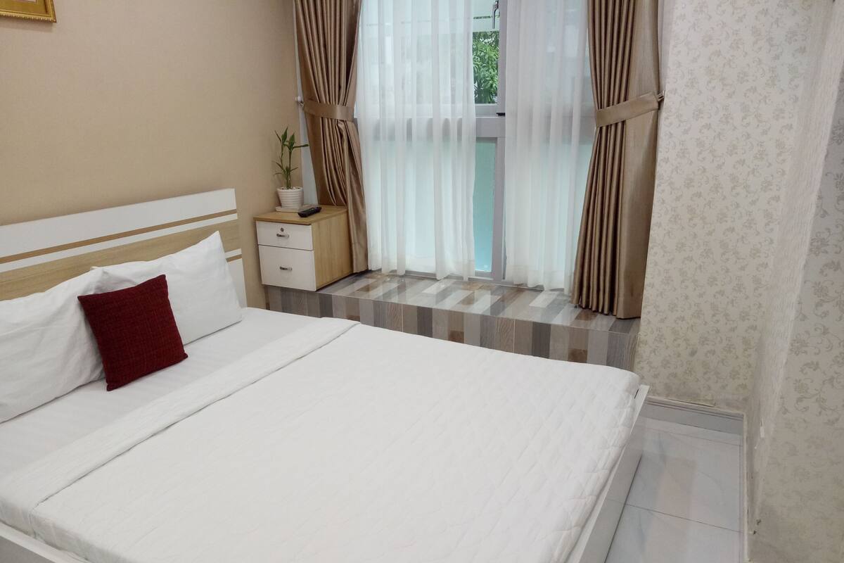 Una cama bien hecha con sábanas blancas y una almohada roja, una mesita de noche con una planta, y una ventana con cortinas.
