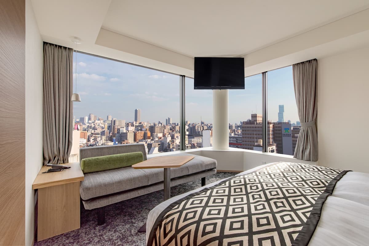 Ein modernes Hotelzimmer mit einem großen Bett, einer Couch, einem kleinen Tisch und einem an der Wand befestigten Fernseher. Das Zimmer verfügt über große Fenster mit Blick auf die Stadt.