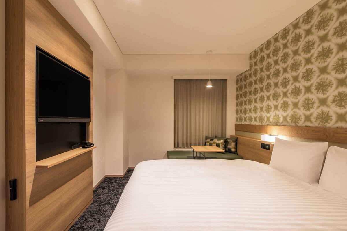 Ein Hotelzimmer mit einem großen Bett, einem flachbildfernseher, der an einer Holzvertäfelung befestigt ist, einem kleinen Tisch und einem Nachttisch mit einer Lampe.