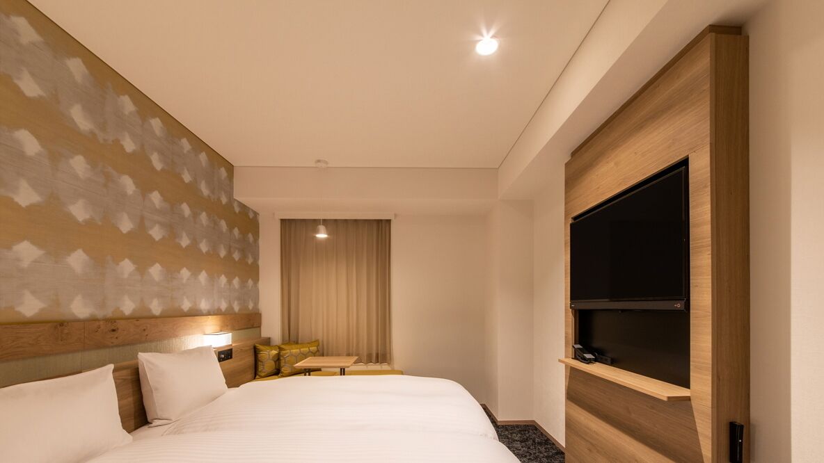 Une chambre d’hôtel avec un grand lit, un téléviseur à écran plat, une tête de lit en bois, une petite table et une fenêtre avec des rideaux.