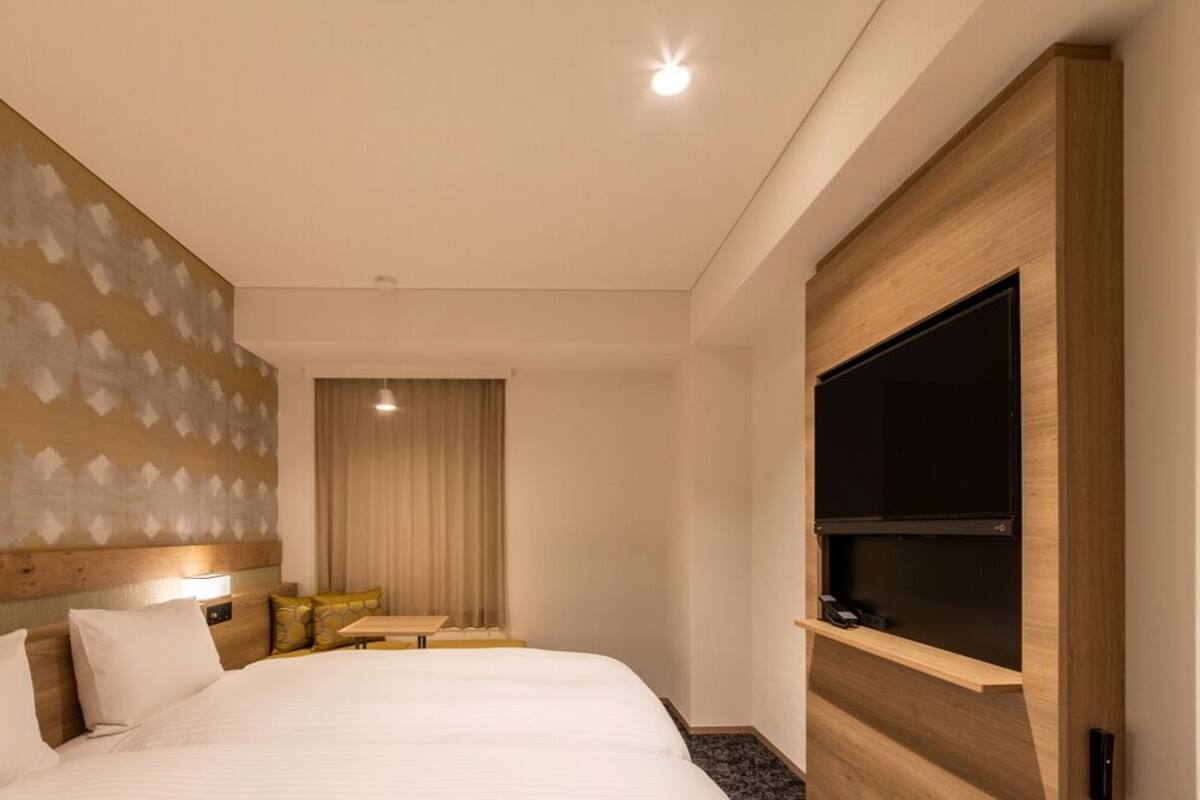 Ein Hotelzimmer mit einem großen Bett, einem Flachbildfernseher, einem Holz-Kopfschild, einem kleinen Tisch und einem Fenster mit Vorhängen.
