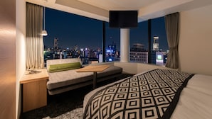 Une chambre d’hôtel dotée d’une grande fenêtre donnant sur la ville, d’un lit avec une couvre-lit à motifs, d’un canapé agrémenté d’un coussin vert et d’une petite table.