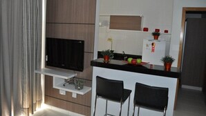 Ein modernes Wohnzimmer mit einem Flachbildfernseher, einer kleinen Sitzecke mit zwei Sesseln, einer Küchenzeile mit Kühlschrank und Topfpflanzen sowie einem großen Fenster mit Vorhängen.