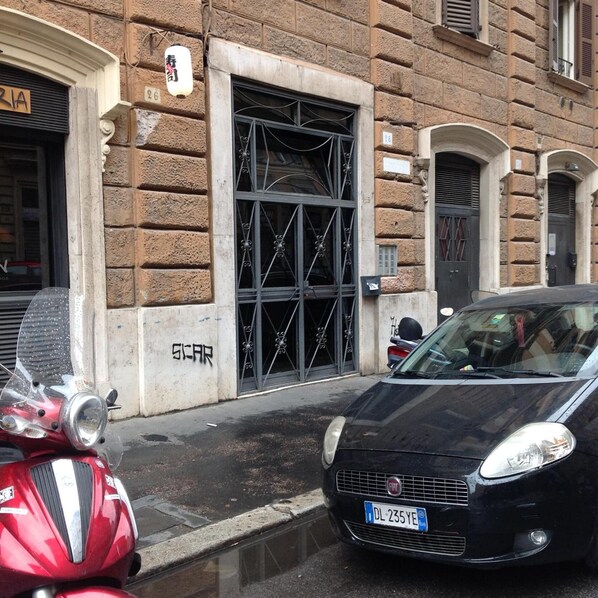A street scene with a parked red scooter, a black car, and a building with a metal gate. - Cardinal Inn (Rome)