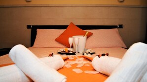 Una cama bien hecha con una colcha rosa y naranja, dos toallas blancas, un plato con aperitivos y una botella de champán.