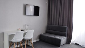 Une chambre d’hôtel moderne avec un canapé gris, un bureau blanc avec deux chaises, un téléviseur à écran plat fixé au mur et une grande fenêtre avec des rideaux.