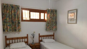 Une chambre avec deux lits simples, une table de chevet avec une lampe, une fenêtre avec des rideaux et un tableau encadré au mur.