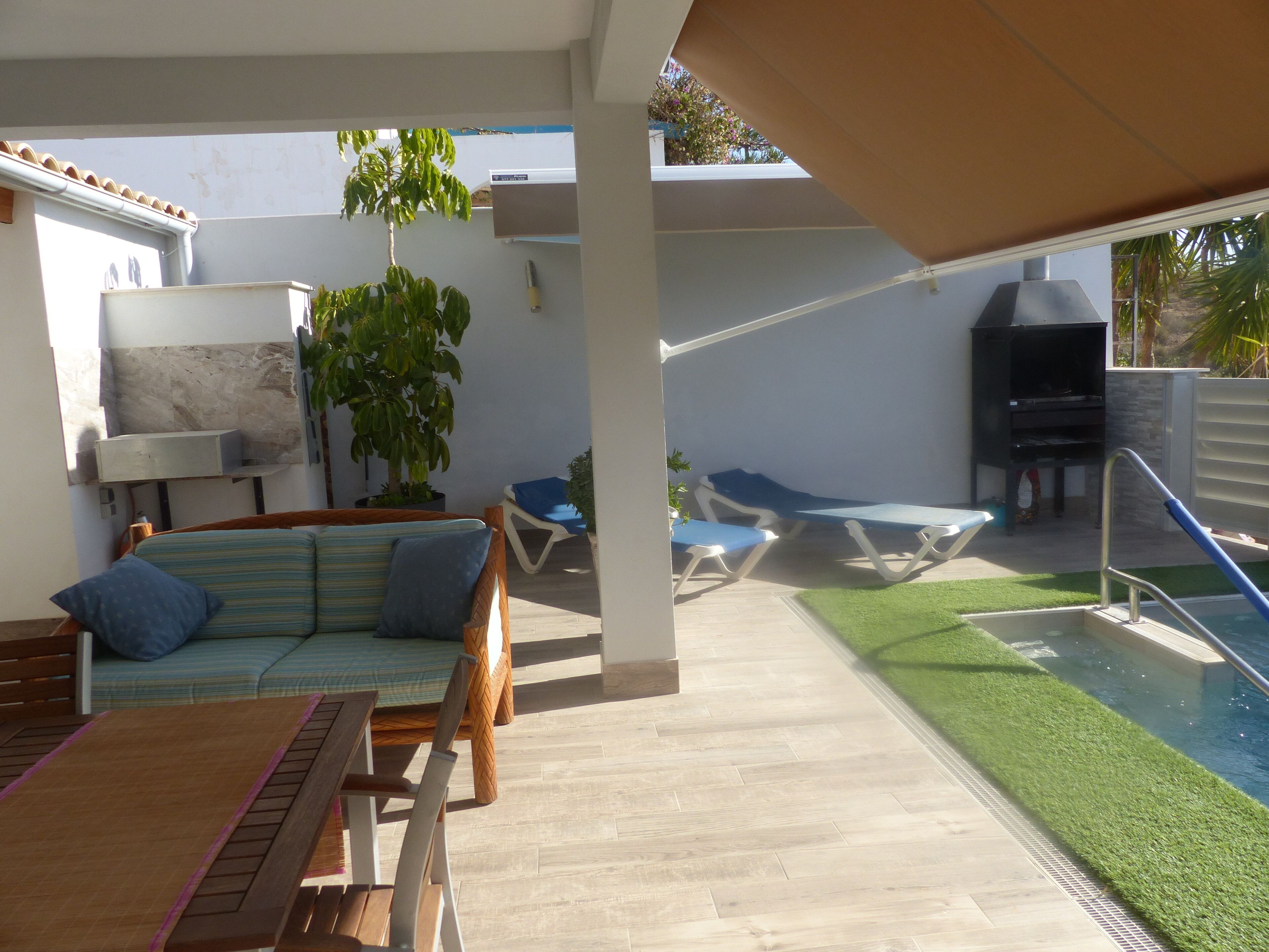 Een terras met een houten tafel, stoelen, een comfortabele bank en een zwembad.