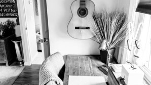 A dining area with a wooden table, a guitar wall art, a potted plant, and a window with blinds.