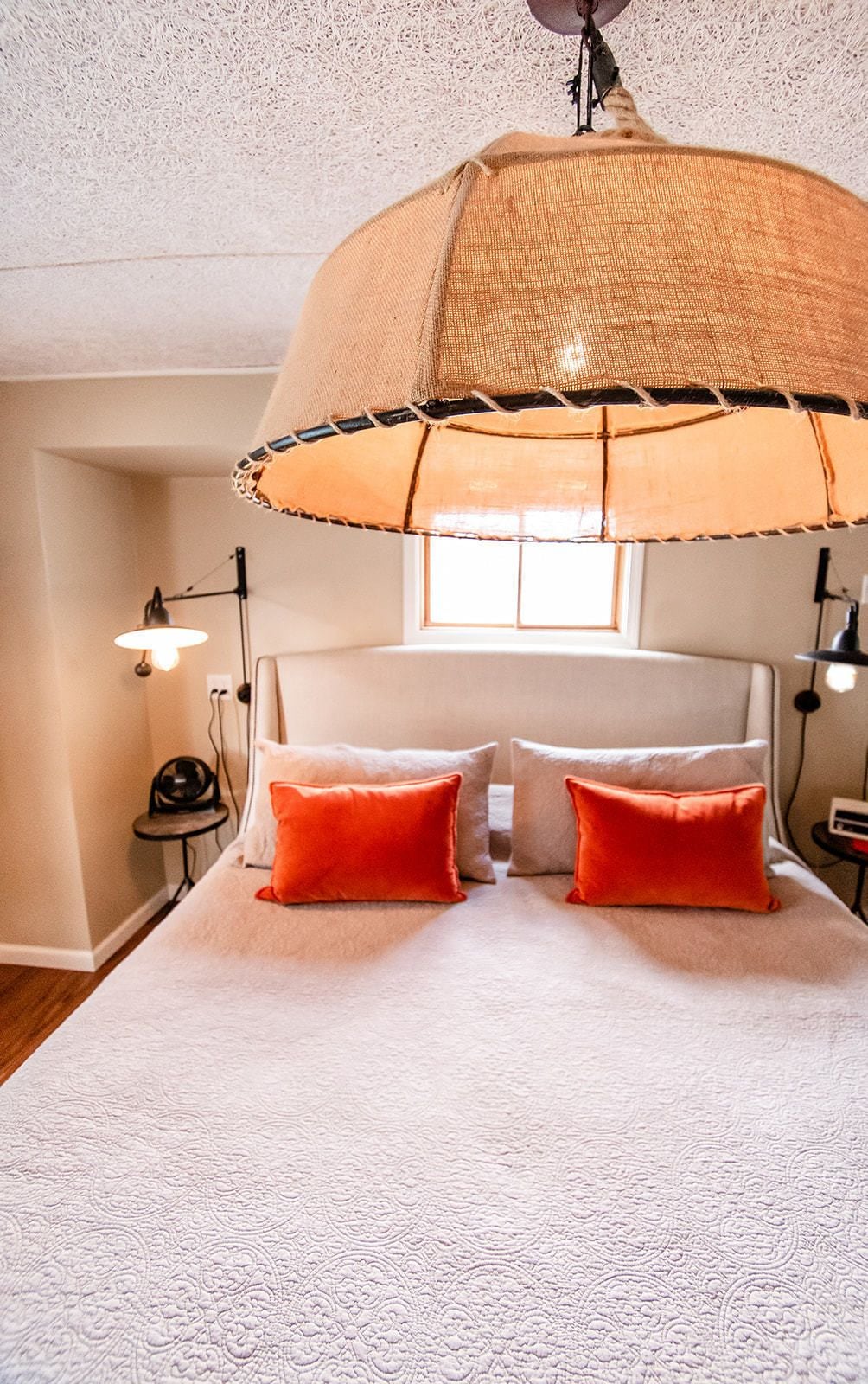 Un dormitorio con una cama grande, dos almohadas rojas, una colcha blanca texturizada y una lámpara colgante con pantalla color beige.