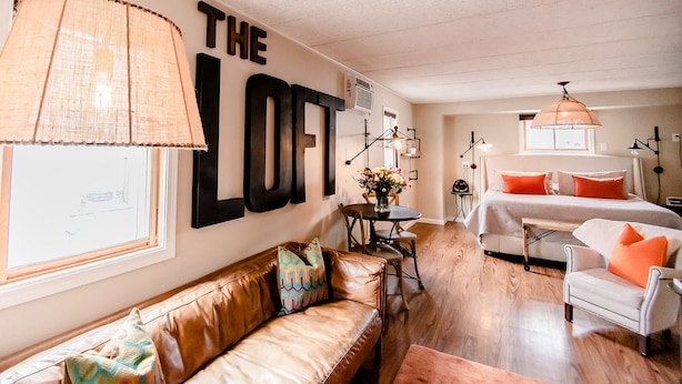 A cozy loft-style room with a large wall-mounted sign, a leather sofa, a dining table with chairs, a bed with orange pillows, and a hanging light fixture.