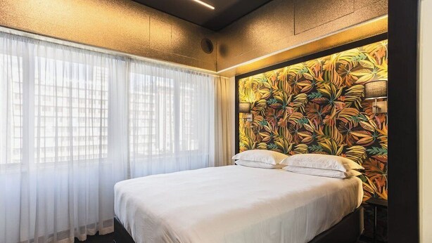 A hotel room with a bed, curtains, and a wall with a leaf pattern.