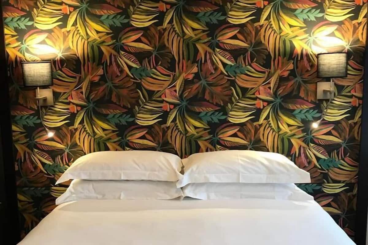 Un lit recouvert de draps blancs et agrémenté d’un plaid marron, placé devant un mur orné d’un motif de feuilles tropicales et éclairé par des lampes fixées au mur.