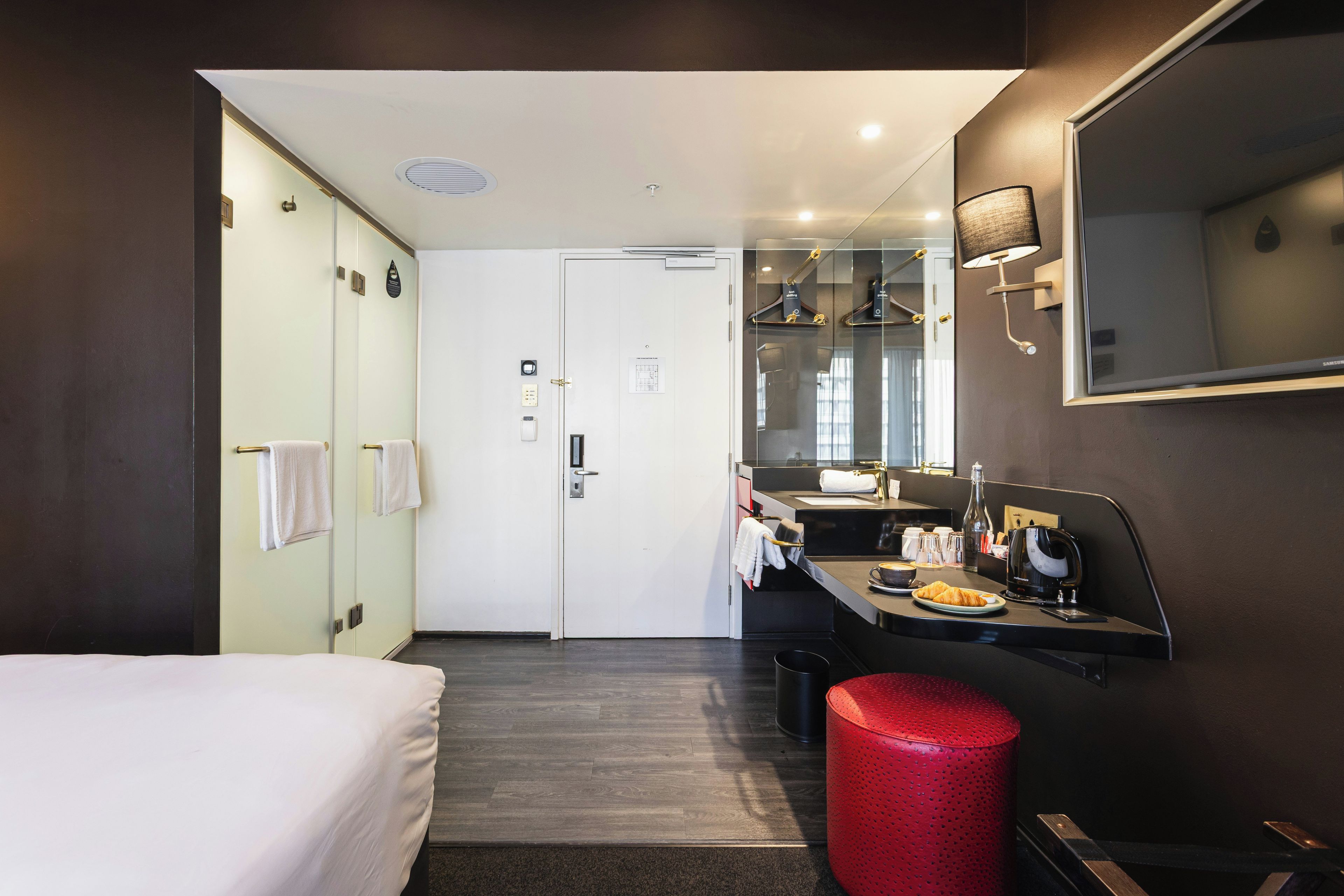 Une chambre d’hôtel avec un lit, une salle de bain dotée d’un miroir et d’un lavabo, ainsi qu’une poubelle rouge.