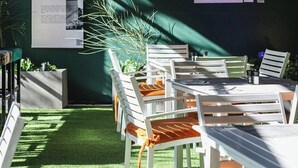 An outdoor seating area with white chairs and tables, a green wall, and a poster of a photographer.
