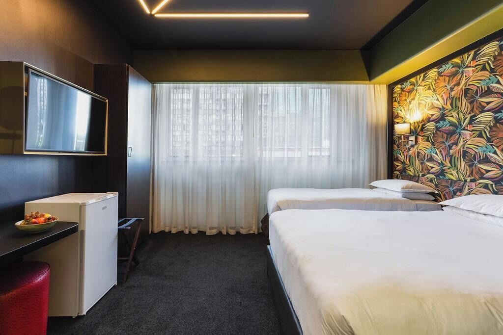 Une chambre d’hôtel comprenant un lit, un bureau avec une télévision, un petit réfrigérateur et une fenêtre avec des rideaux.