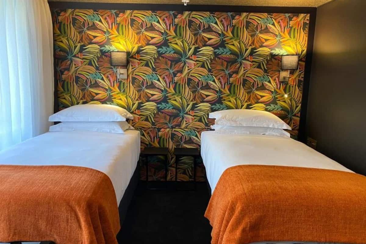 Une chambre d’hôtel avec deux lits simples, une tête de lit ornée d’un motif de feuilles tropicales et une petite table avec une lampe.