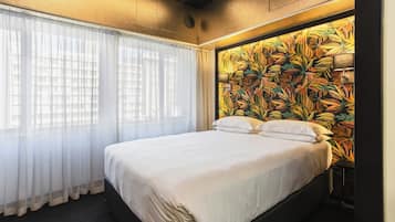 Une chambre d’hôtel avec un grand lit, des rideaux légers et transparents, une tête de lit à motifs floraux et une fenêtre bien visible.