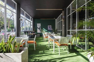 Espaço moderno em ambiente interno com parede verde, grandes janelas e área de estar com cadeiras brancas e almofadas laranja.