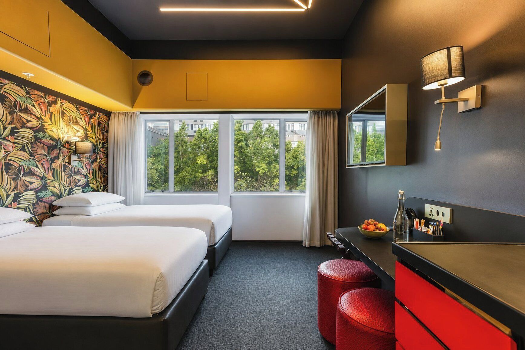 Une chambre d’hôtel avec deux lits, une grande fenêtre, un banc rouge, un bureau avec un miroir et un mur orné d’un motif de feuilles tropicales.