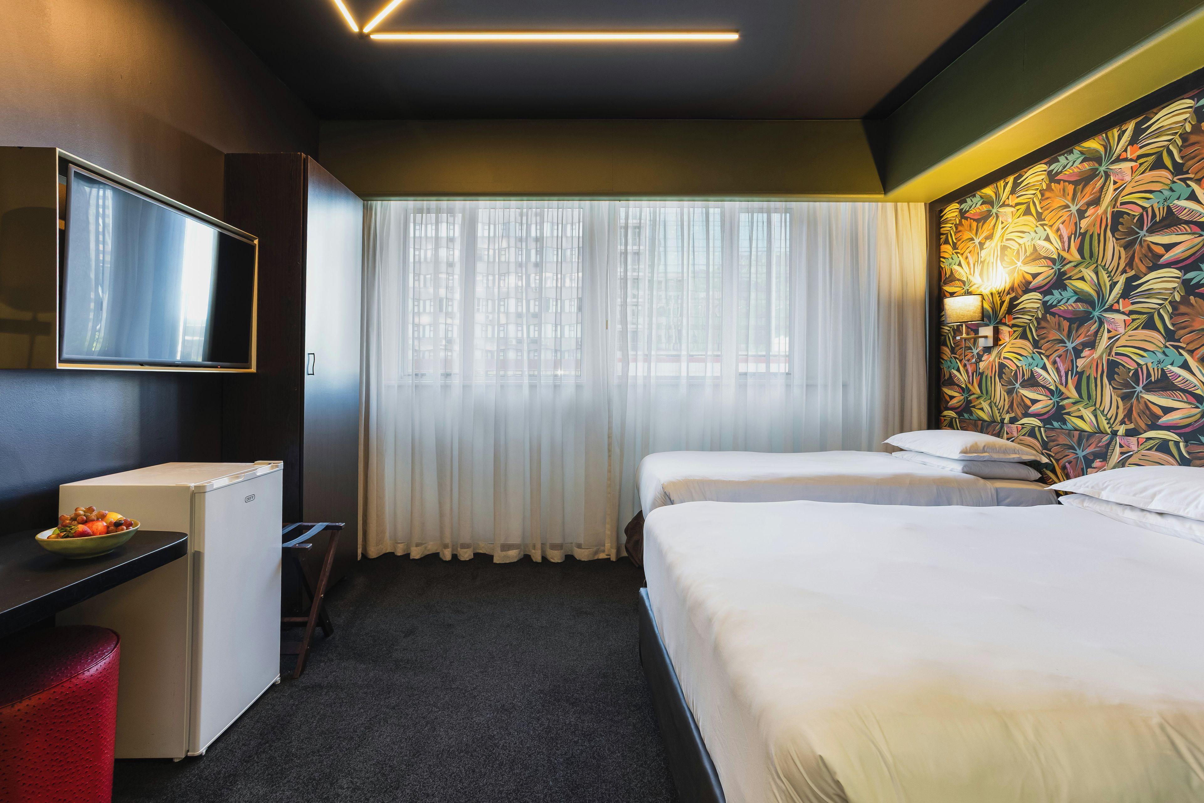Une chambre d’hôtel avec un lit, un bureau avec une télévision, un réfrigérateur et une fenêtre avec des rideaux légers.
