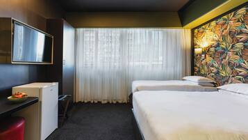 Une chambre d’hôtel avec un lit, un bureau avec une télévision, un réfrigérateur et une fenêtre avec des rideaux légers.