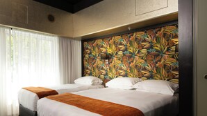A hotel room with two beds, white bedding, a tropical leaf-patterned headboard, and a window with sheer curtains.