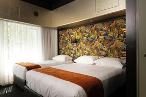 A hotel room with two beds, white bedding, a tropical leaf-patterned headboard, and a window with sheer curtains.